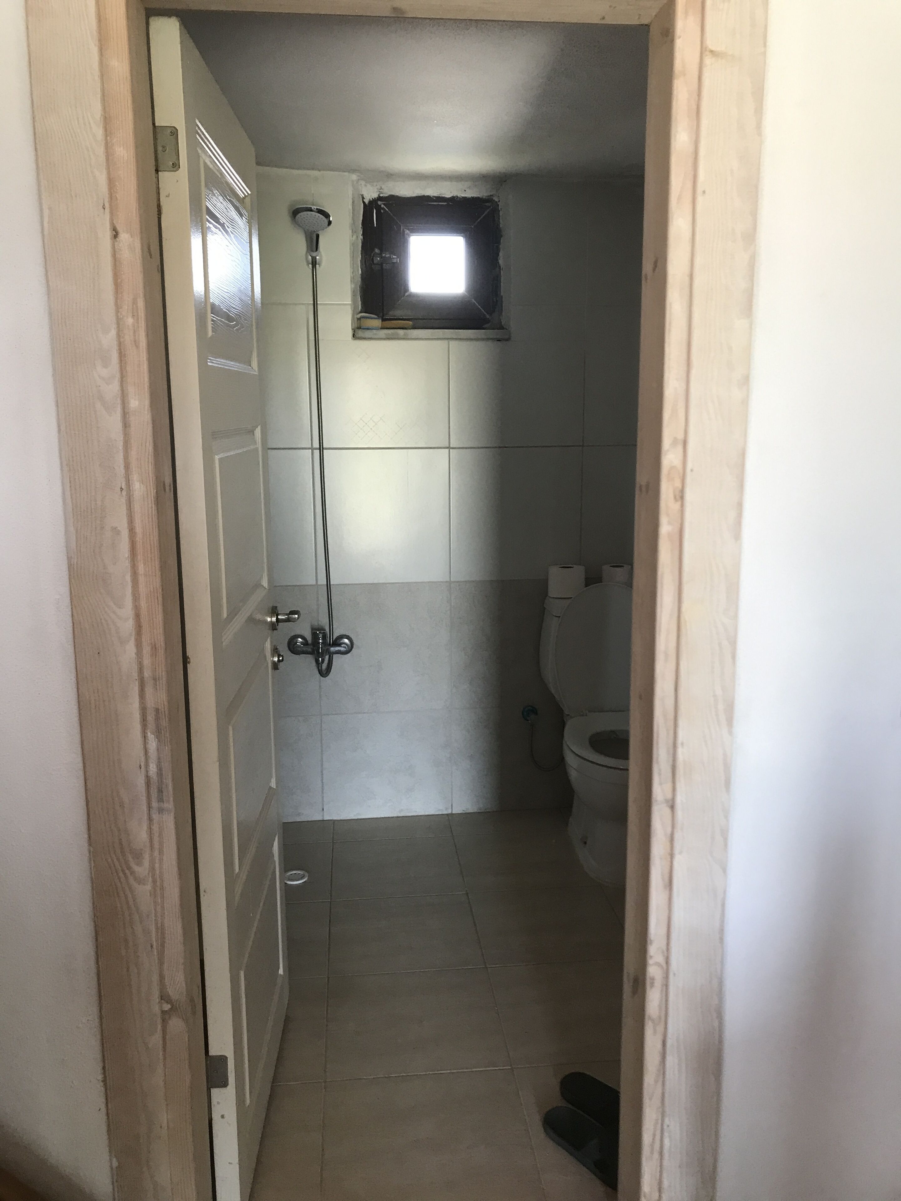 Een smalle badkamer met een douche, toilet en een klein raam.