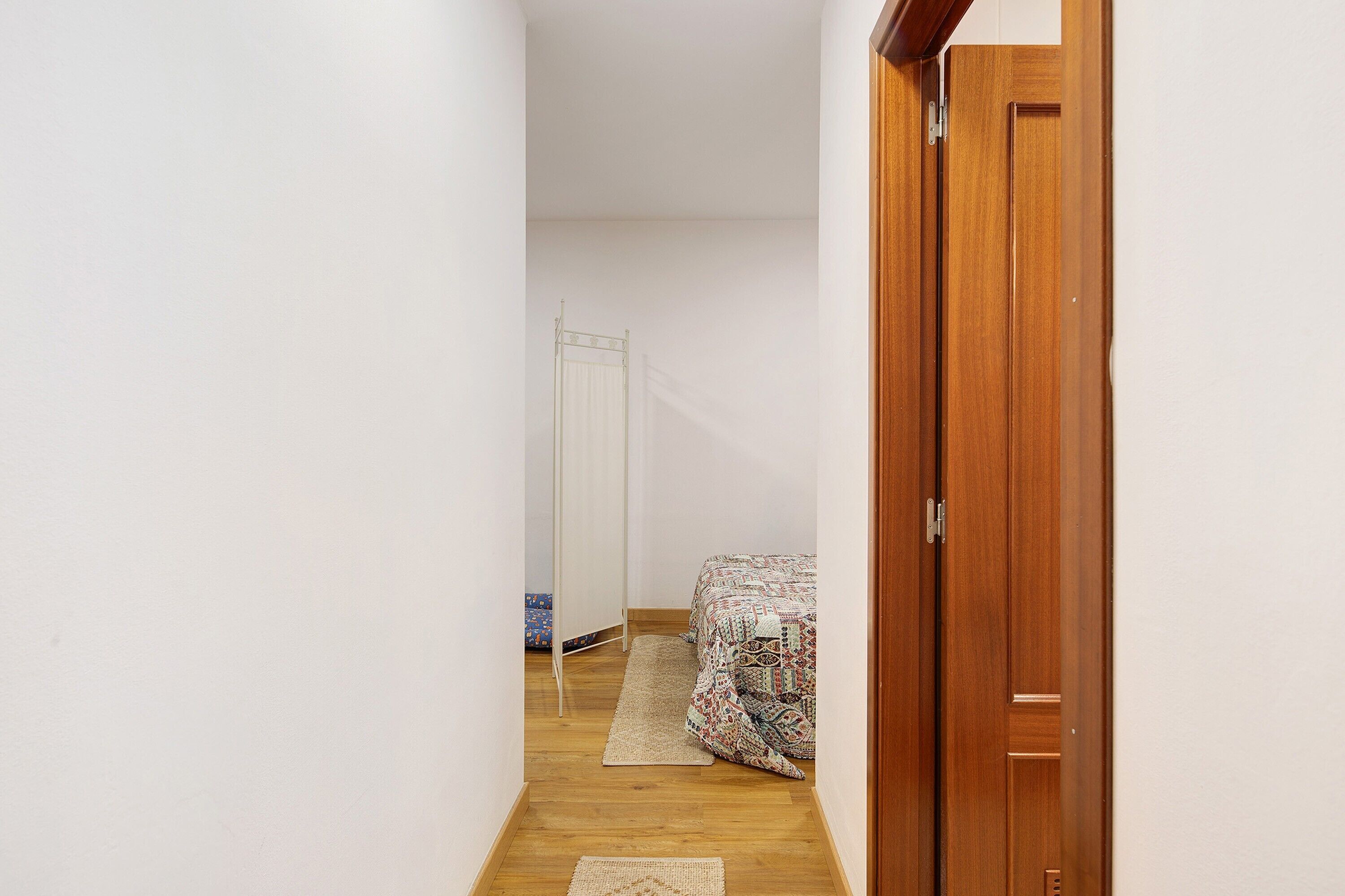 Un couloir étroit avec une porte en bois sur la droite, un lit avec une literie à motifs, une armoire blanche et un petit tapis sur le sol.