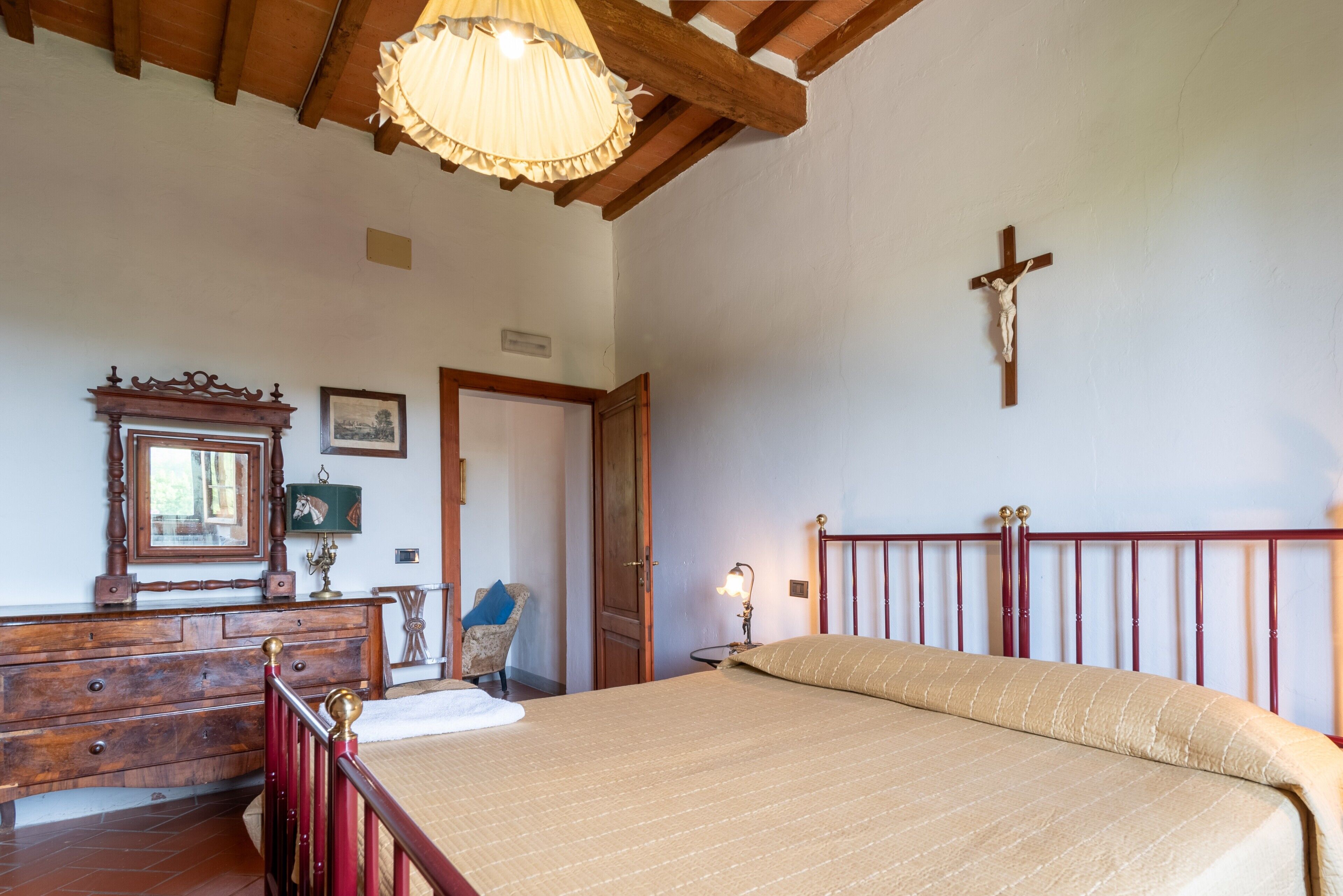 A bedroom with a wooden bed, a wooden dresser, a chair, a cross on the wall, and a door.