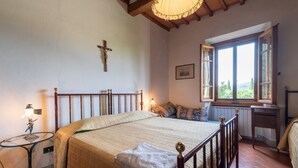 A bedroom with a bed, a cross on the wall, a window with a view, a bedside table with a lamp, and a radiator. - Farmhouse "Fattoria" with Shared Pool, Private Garden & Wi-Fi (Laterina)