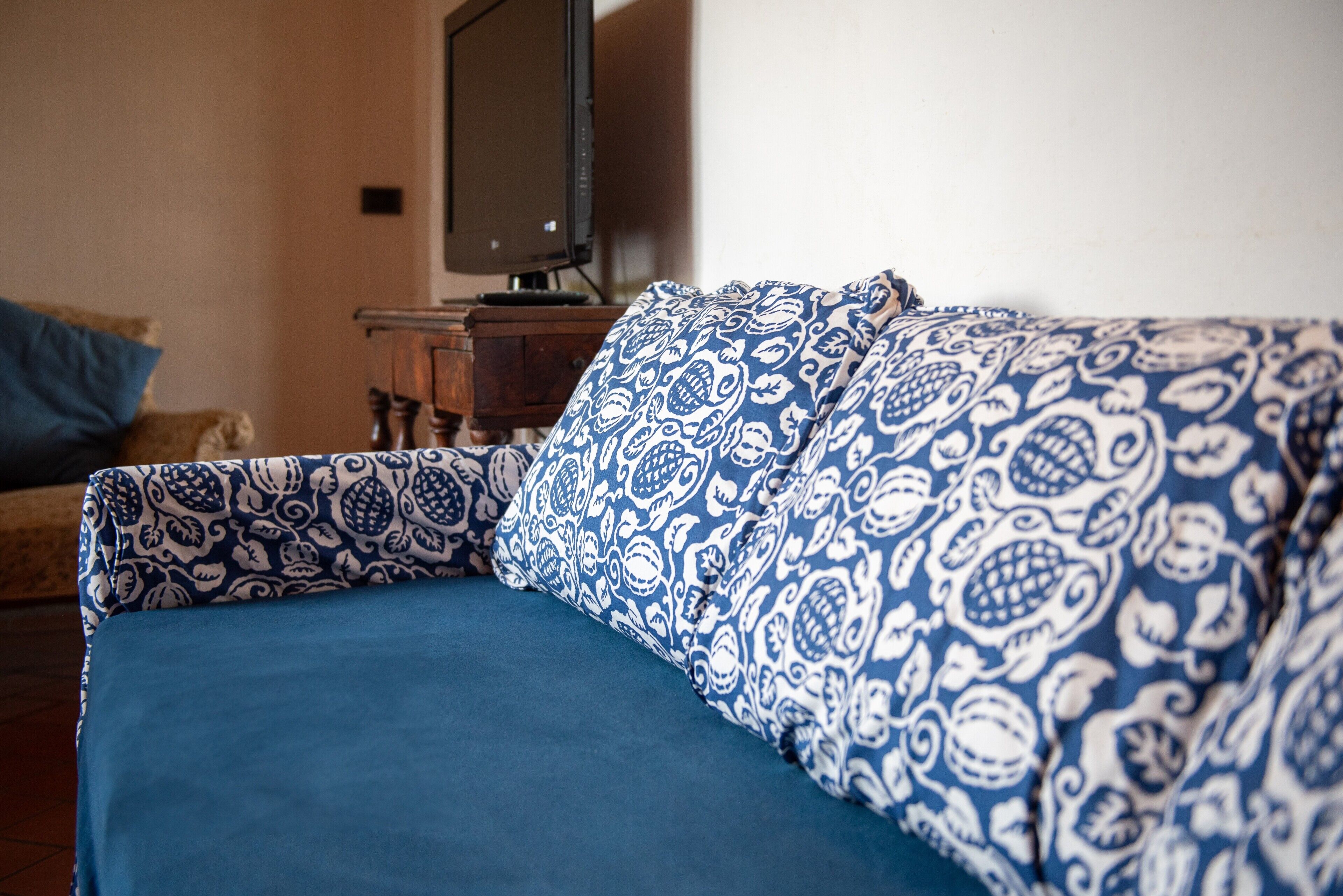 A bed with a patterned blue and white cover, a television on a wooden stand, and a wall with a light switch.
