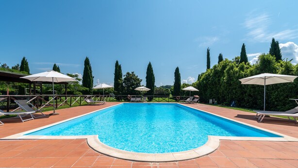 A rectangular swimming pool with a tiled deck, surrounded by lounge chairs and umbrellas, and tall trees in the background. - Farmhouse 'Mulino ' with Shared Pool, Terrace & Wi-Fi (Laterina)