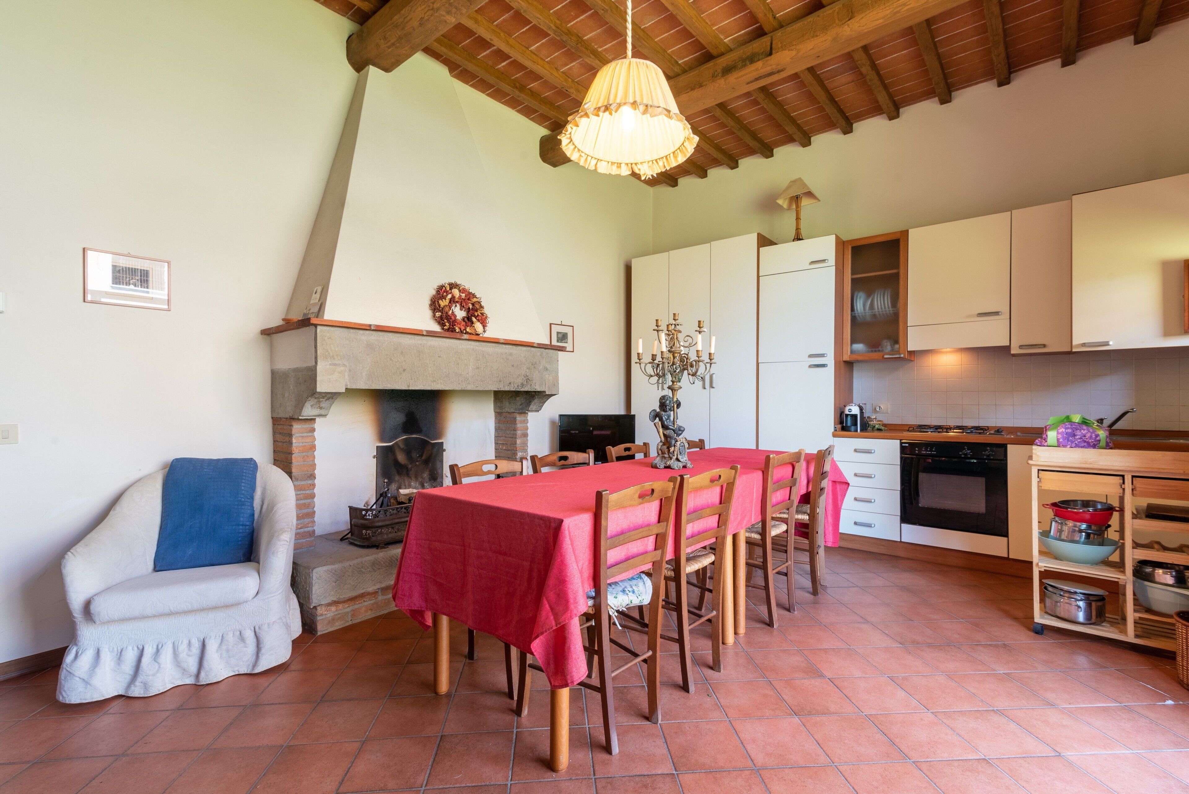 A dining area with a table set for six, wooden chairs, a fireplace, a chandelier, and a kitchen with cabinets and appliances.
