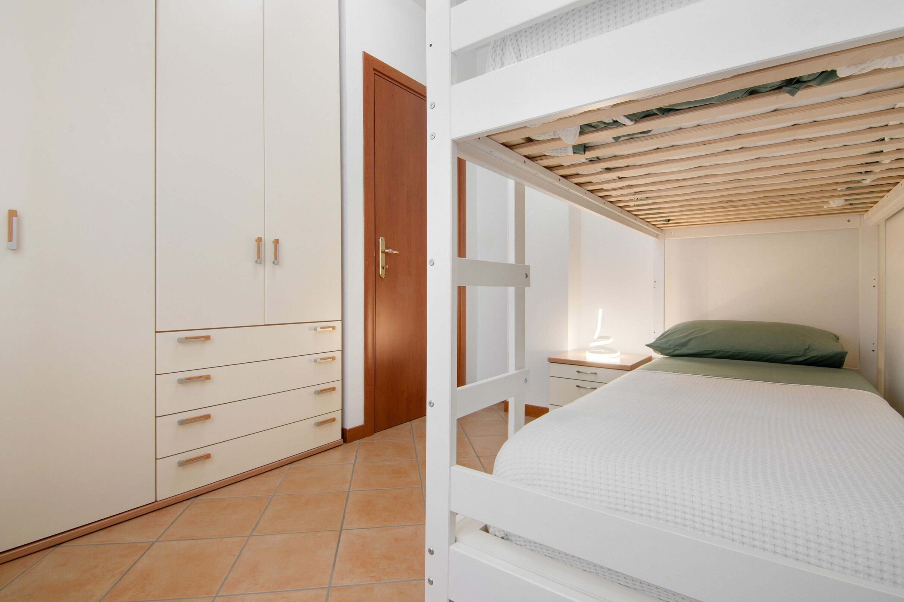 A bedroom with a bunk bed, a wooden door, a white wardrobe, and a bedside table.