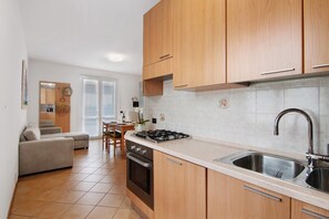 A modern kitchen with wooden cabinets, a gas stove, a sink, and a dining area with a chair and table. - Casa Enea with private garden and mini pool (Colico)