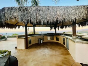 An outdoor kitchen area with a thatched roof, a built-in grill, and a view of the landscape.