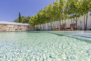 Una piscina cristalina con sillones de madera y un muro de piedra al fondo.
