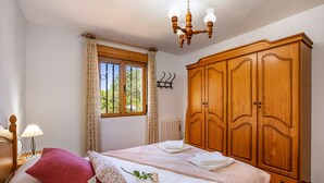 A bedroom with a bed, a wooden wardrobe, a window with curtains, a radiator, and a chandelier. - Holiday Home 'Ladera De Los Molinos' with Mountain View, Pool, Garden & Wi-Fi (Padul)