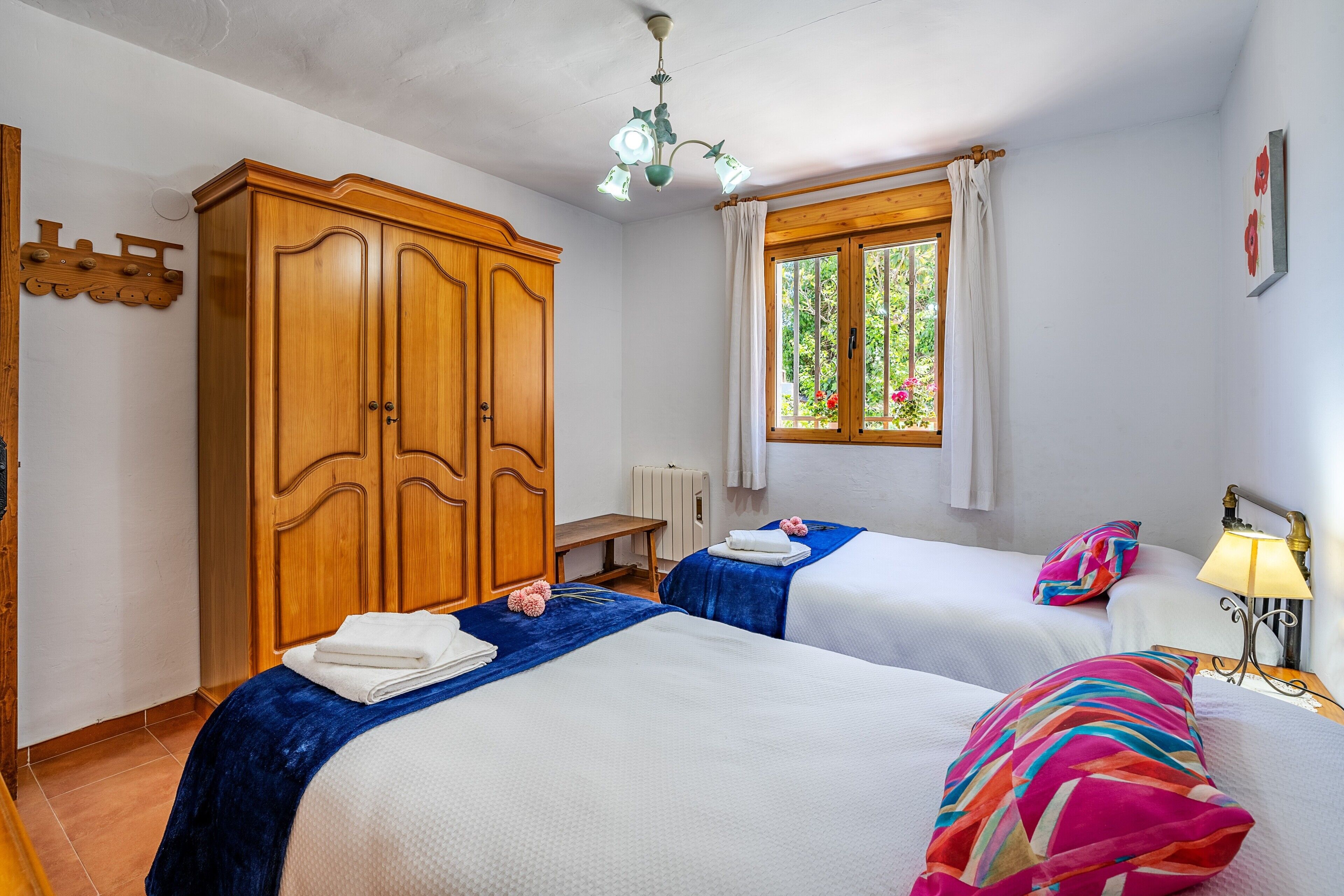 Una camera d'albergo con due letti, un armadio in legno, una finestra con tende, un termosifone e una lampada da comodino.