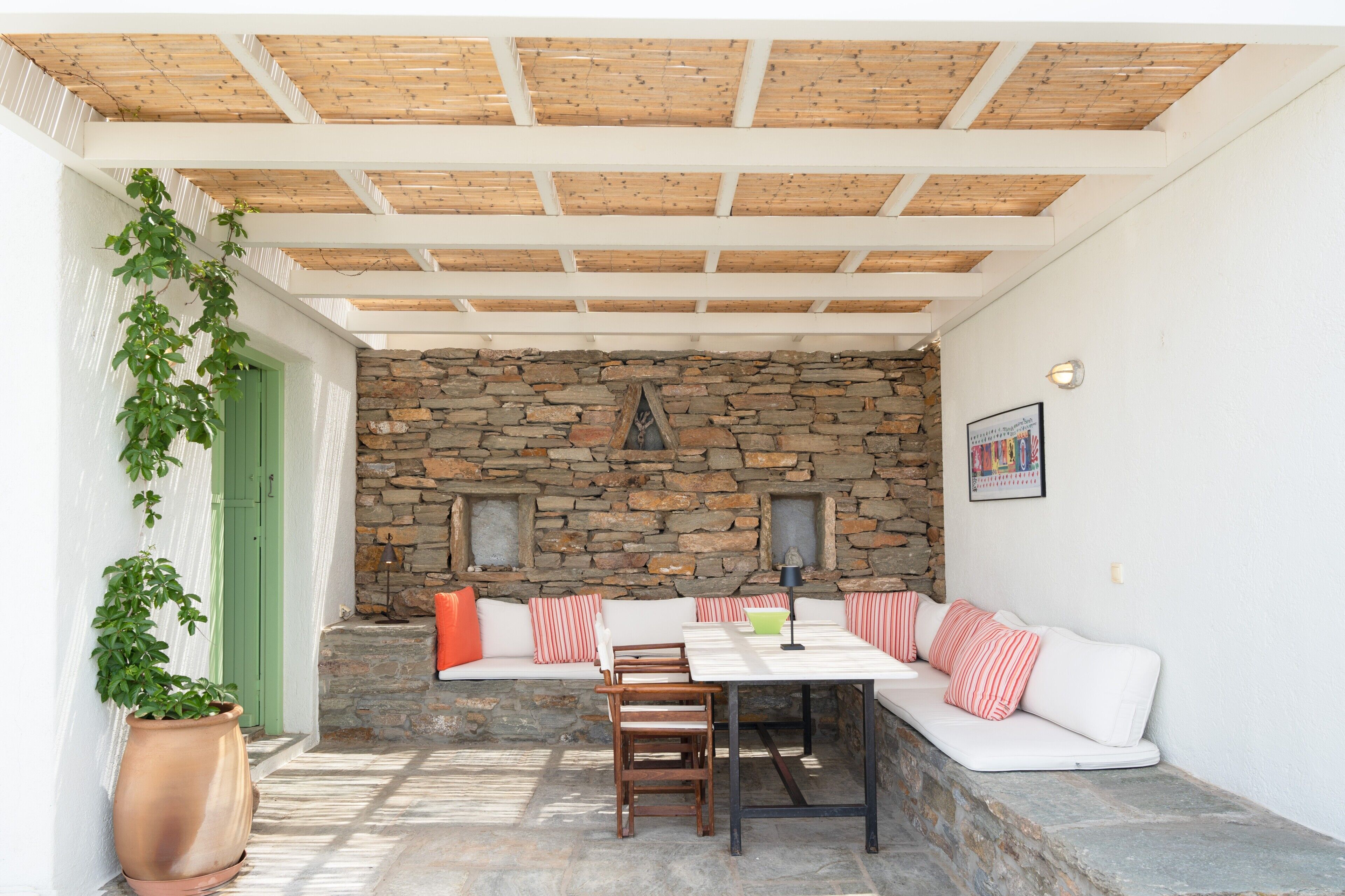 A covered patio with a stone wall, wooden ceiling, and a seating area with cushions.