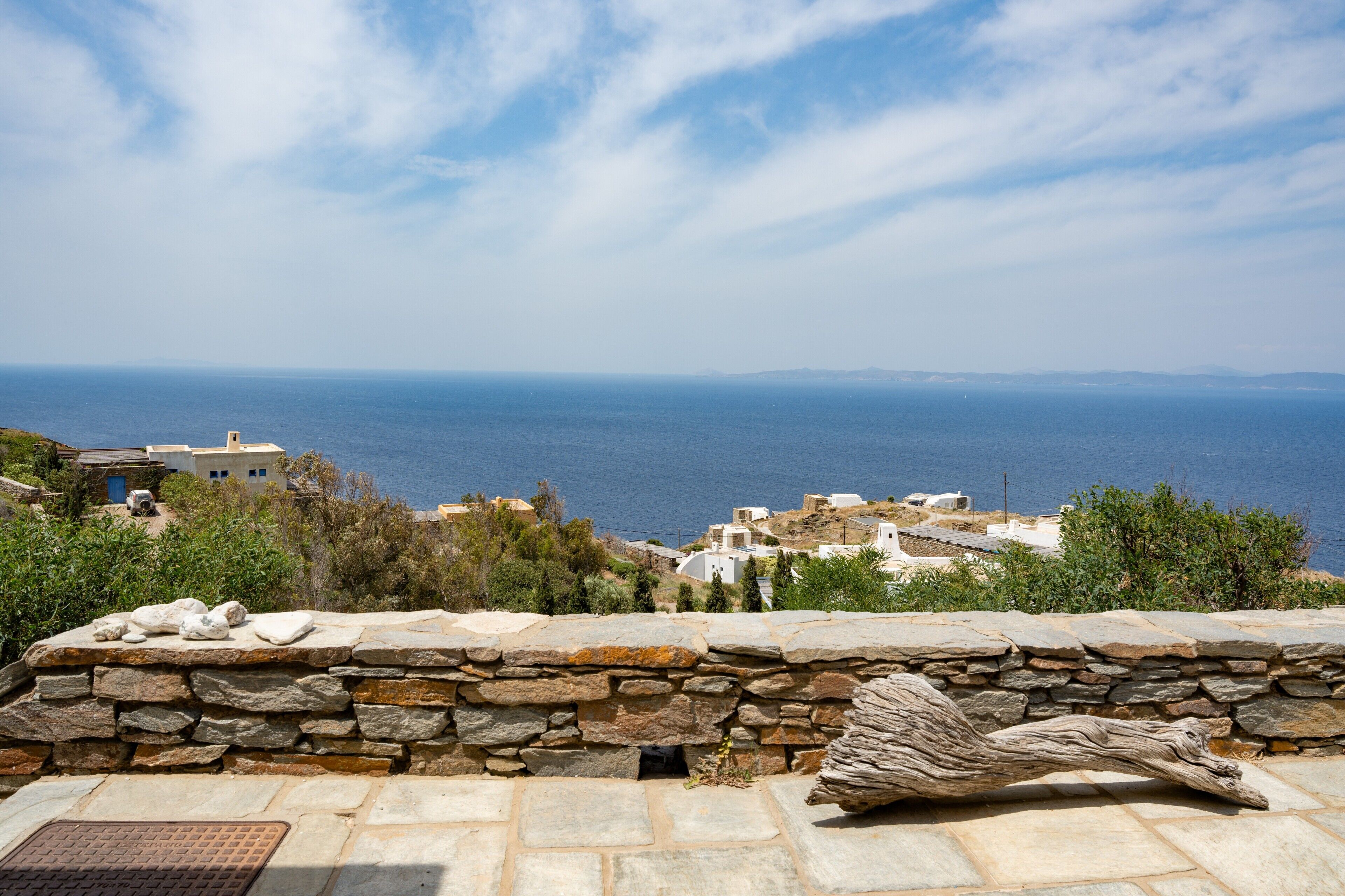 A stone wall with a rustic wooden sculpture, a clear view of the sea, and buildings in the background.