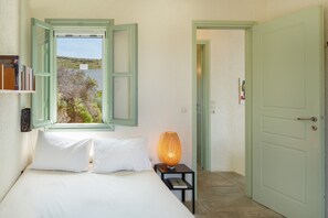 Un dormitorio con cama, una mesita de noche con lámpara, una ventana con contraventanas abiertas y una puerta.