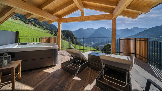 Een houten terras met een jacuzzi, rieten stoelen en uitzicht op bergen.