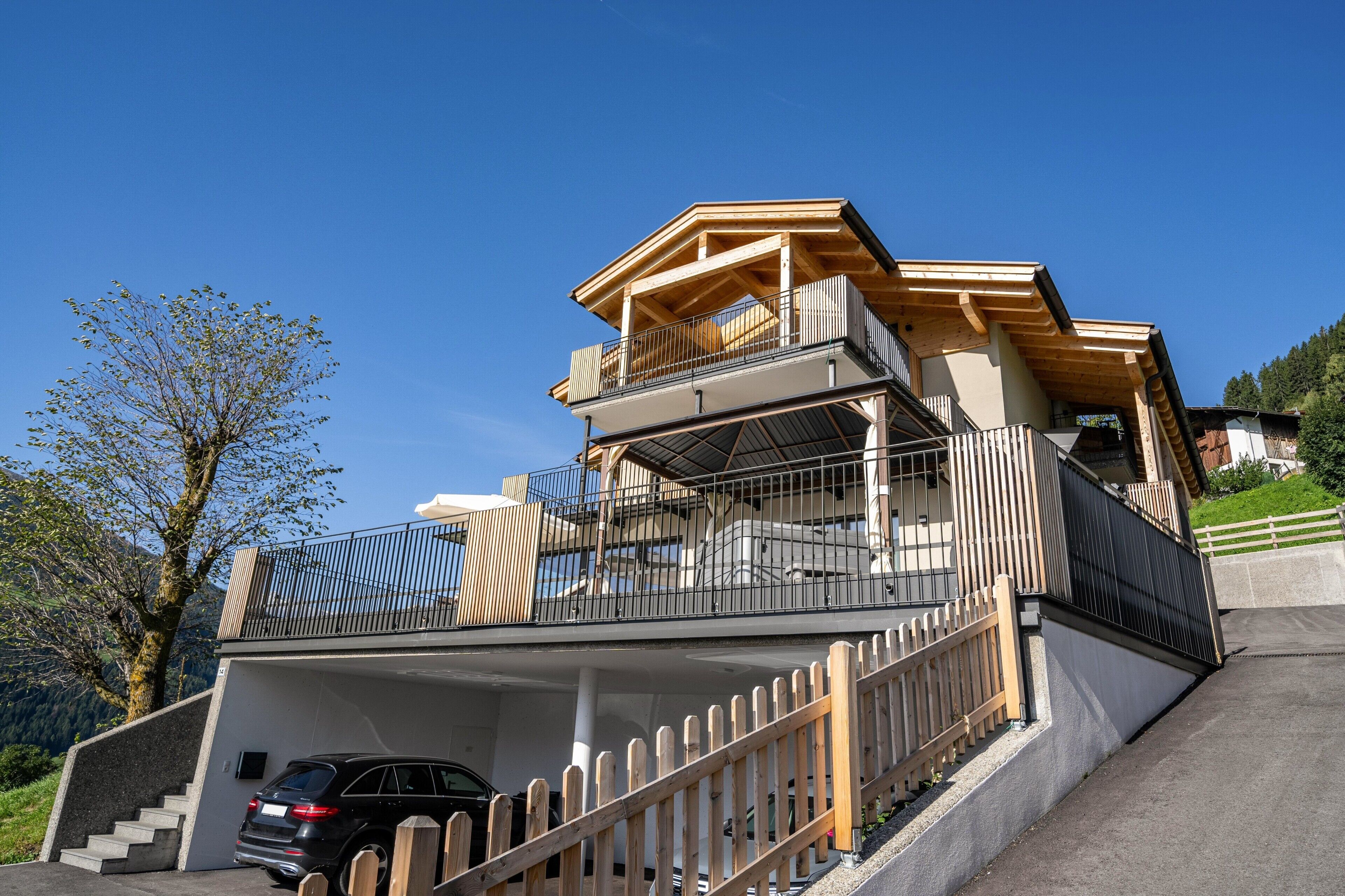 Une maison à plusieurs niveaux avec une voiture garée dans l’allée, des rampes en bois et un ciel bleu dégagé.