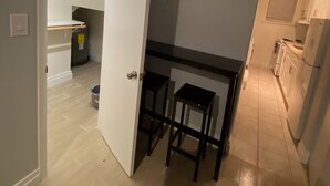 Una cucina con un piano di lavoro nero e due sgabelli, una porta bianca che dà su una scala, e un pavimento chiaro.