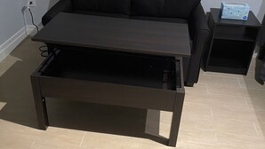 Un soggiorno moderno con un divano nero, un tavolino in legno con cassetto e un piccolo tavolino laterale con una scatola sopra.