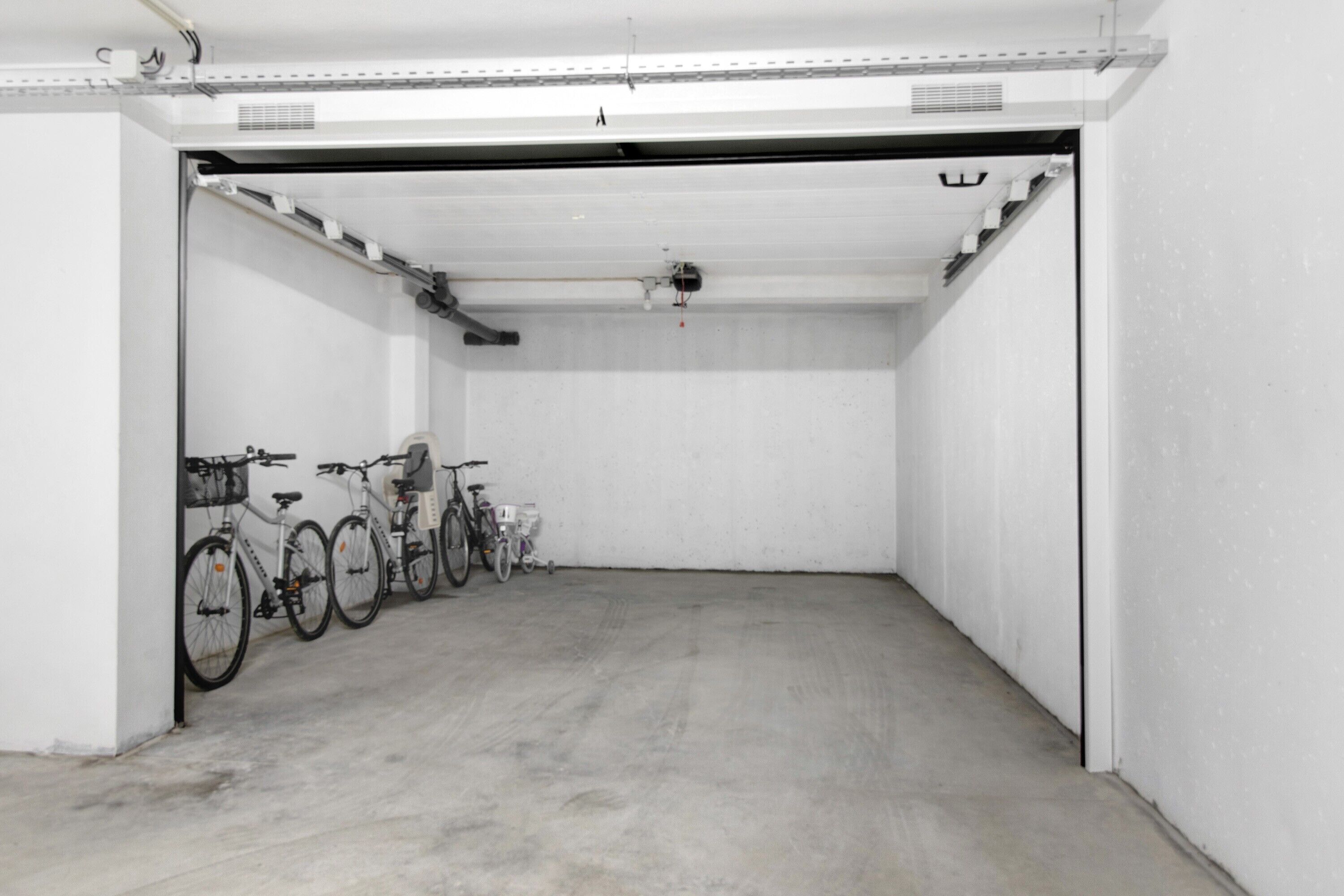 Un ambiente interno pulito e vuoto, con pavimento in cemento, pareti bianche e una fila di biciclette appoggiate a una parete.