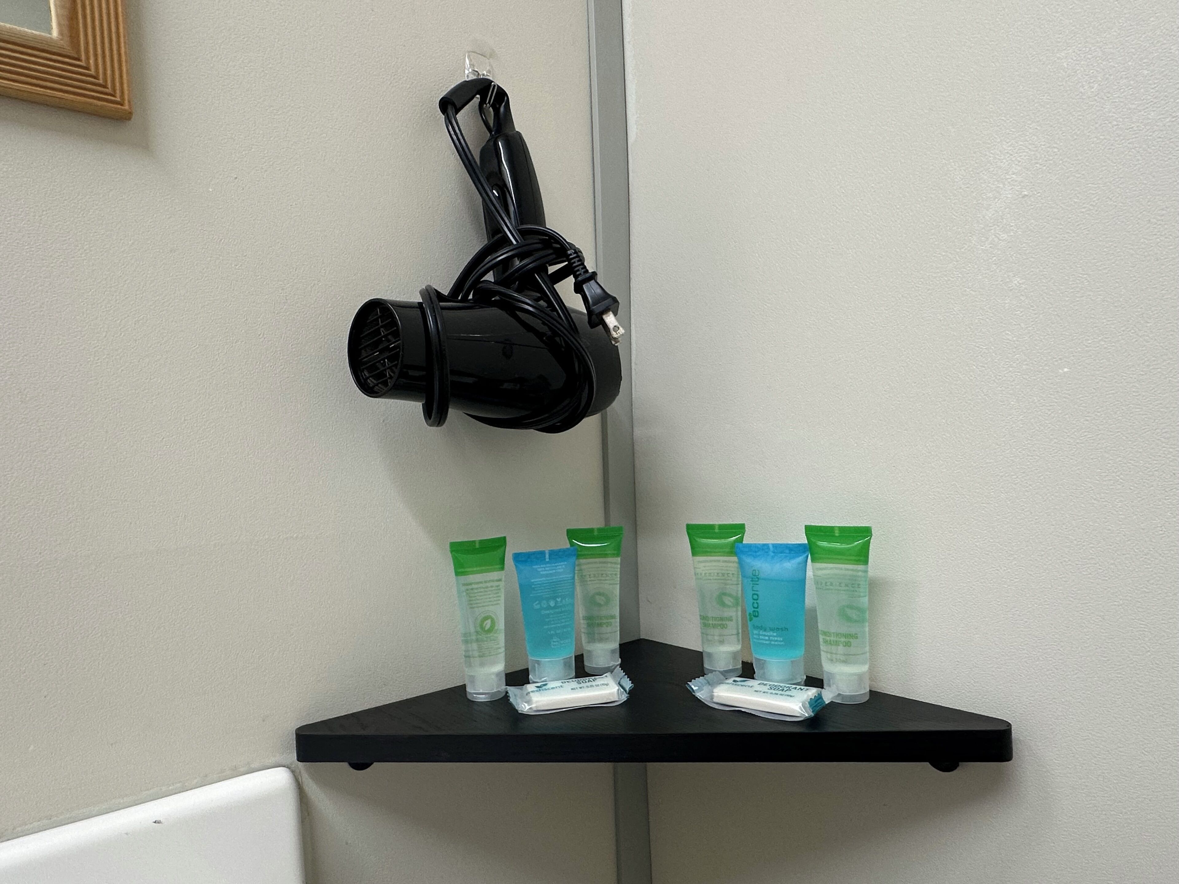 A black hair dryer hanging on a wall, a black shelf with green and blue shampoo bottles, and a white wall.