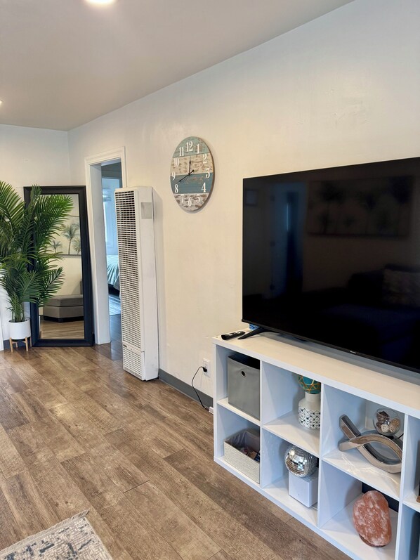 A modern living room with a flat-screen TV on a white stand, a decorative clock, and a potted plant. - Casa Bella (Seaside)