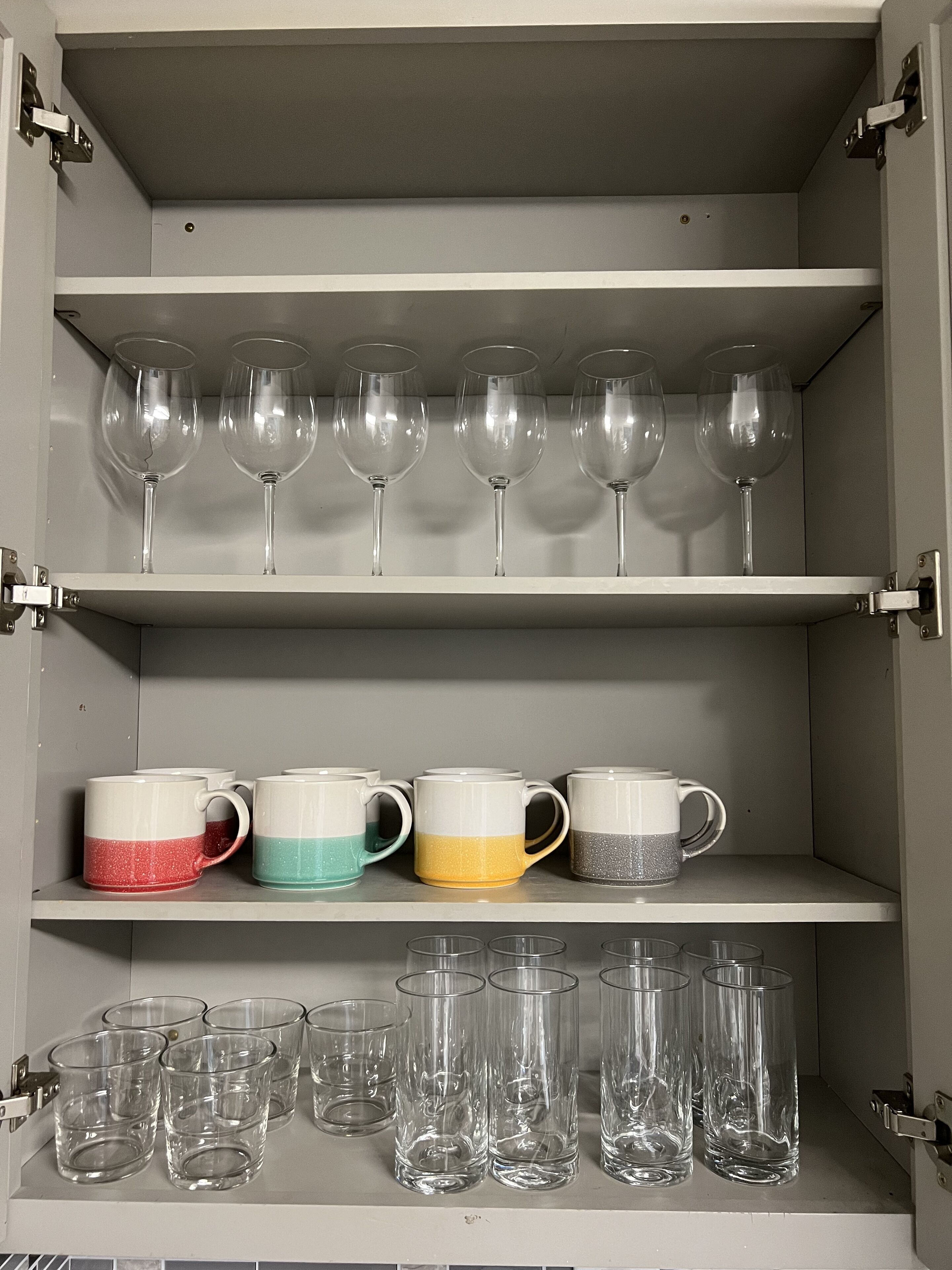 A storage cabinet with shelves holding wine glasses, ceramic mugs, and clear drinking glasses.