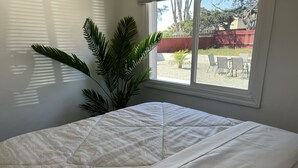 Un letto rifatto con copriletto bianco, una pianta in vaso accanto a una finestra e vista su un patio esterno.
