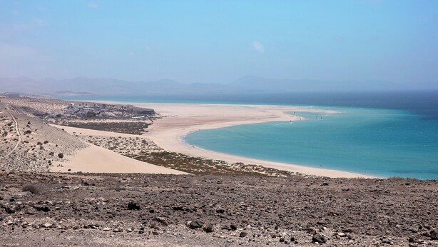 A coastal landscape with a beach, rocky terrain, and distant mountains.