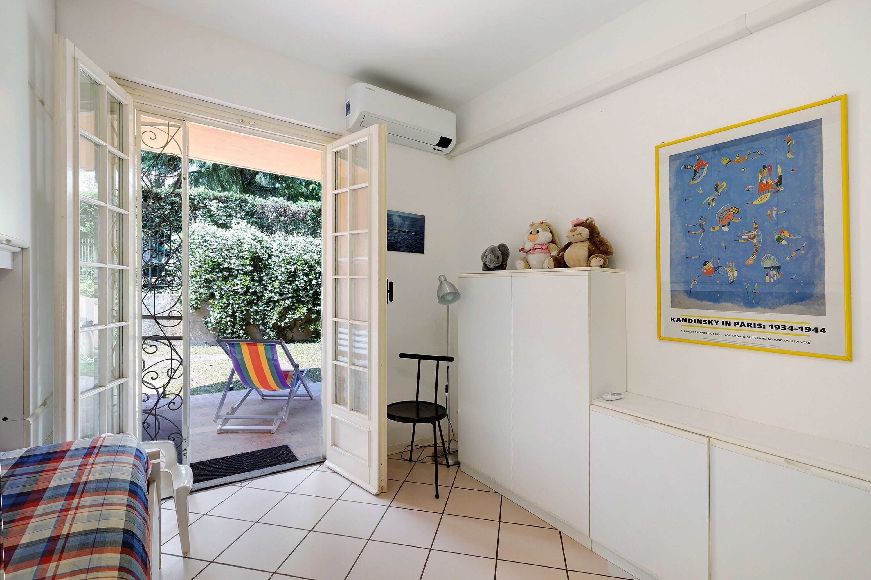 Une pièce comprenant un lit, une chaise, une petite table, un tableau encadré accroché au mur et une porte donnant sur un espace extérieur avec une chaise et une table.