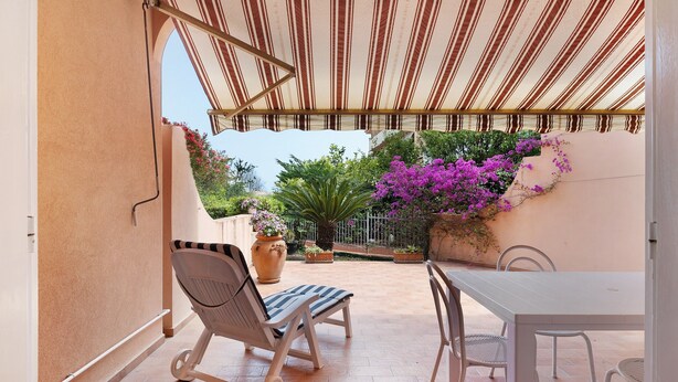 Eine überdachte Terrasse mit Tisch und Stühlen, einem Blumentopf und Blick auf den Garten.