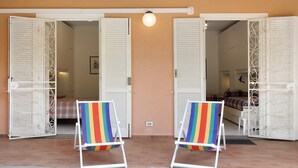 Ein Zimmer mit zwei geöffneten weißen Türen, die in einen anderen Raum führen, einem Bett mit karierter Decke und zwei bunten Stühlen.