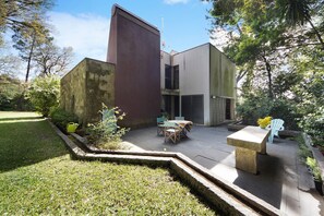 A modern outdoor patio with a table and chairs, surrounded by greenery and a stone wall.