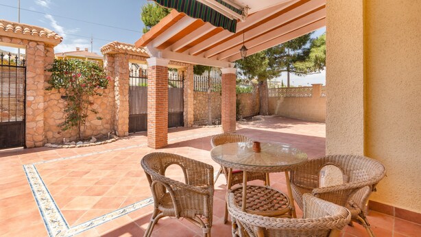 A covered patio with wicker furniture, a glass table, and a stone wall.