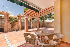 Une terrasse couverte, aménagée avec des meubles en osier, une table en verre et un mur en pierre.