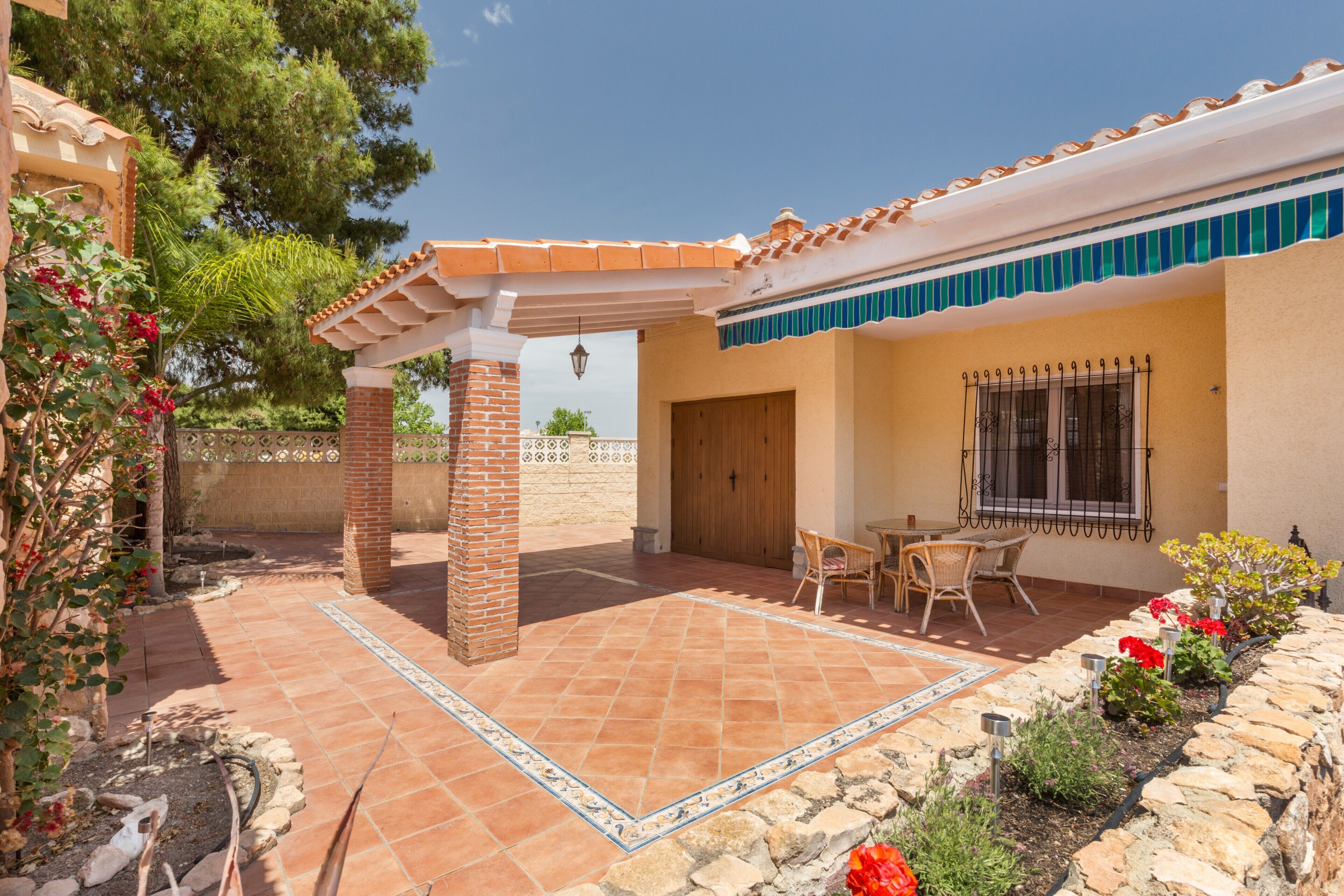 Un patio con suelo de baldosas, una pérgola, un muro de piedra y una ventana con reja de metal.