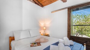 Una cama bien hecha con una revista, un sombrero y toallas, una mesita de noche con una lámpara, y una ventana con vistas a árboles.