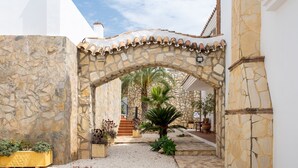 Un arco de piedra que da a un patio con una palmera, plantas en macetas y una escalera.