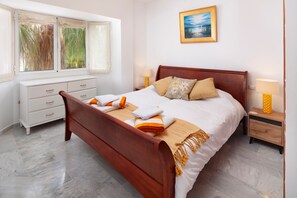 Een netjes opgemaakt bed met een houten hoofdbord, een witte nachtkastje met een lamp, een raam met uitzicht op groen, en een schilderij aan de muur.