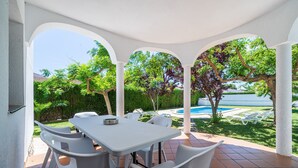 Un patio techado con mesa y sillas, una piscina al fondo y follaje.