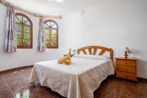 A bedroom with a bed, a nightstand, two windows with curtains, and a tiled floor.