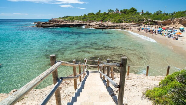 Una playa con agua cristalina, una escalera de madera que baja hacia la orilla y una hilera de sombrillas en la arena.