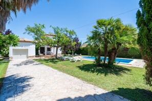 A stone pathway leading to a white building with a garage, a swimming pool, and palm trees.
