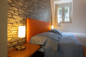 Une chambre avec un mur en pierre, une table de chevet en bois avec une lampe, un lit recouvert d’une couverture à rayures et une fenêtre avec des rideaux.