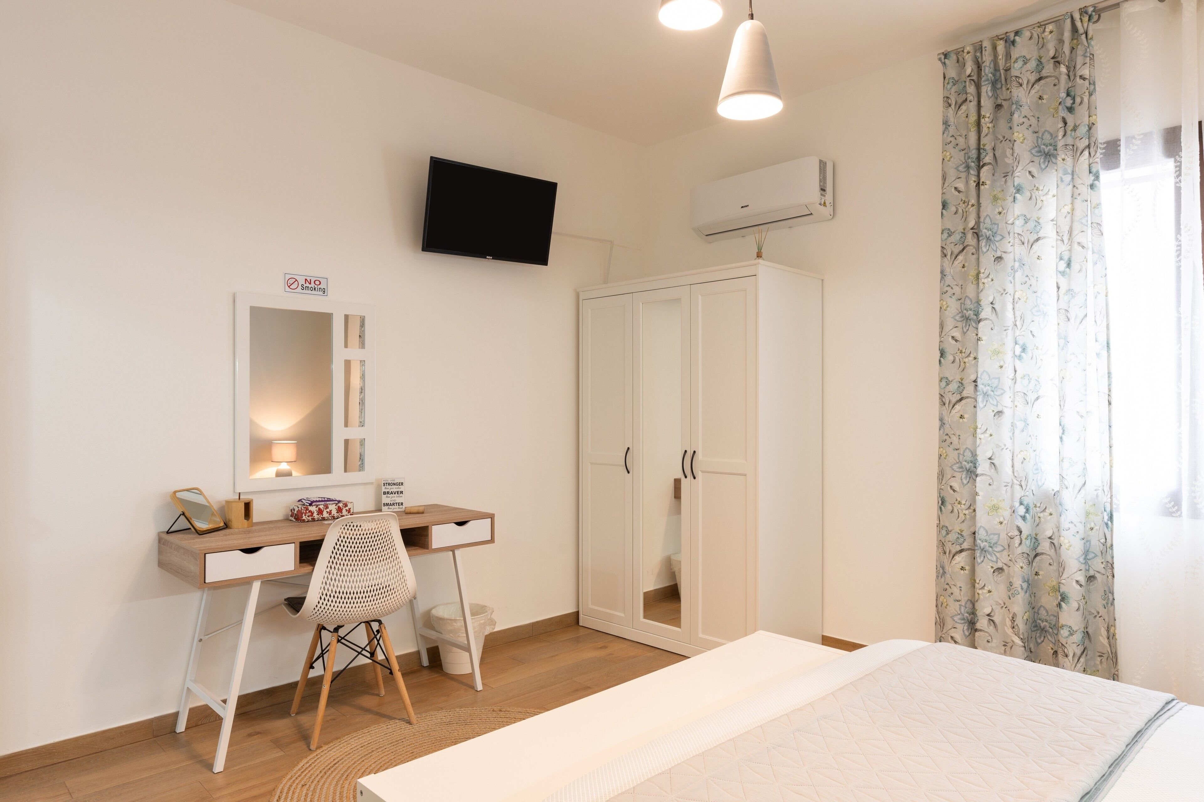 Ein Schlafzimmer mit Bett, Schreibtisch, Stuhl, Kleiderschrank, Fernseher und einem Fenster mit Vorhängen.