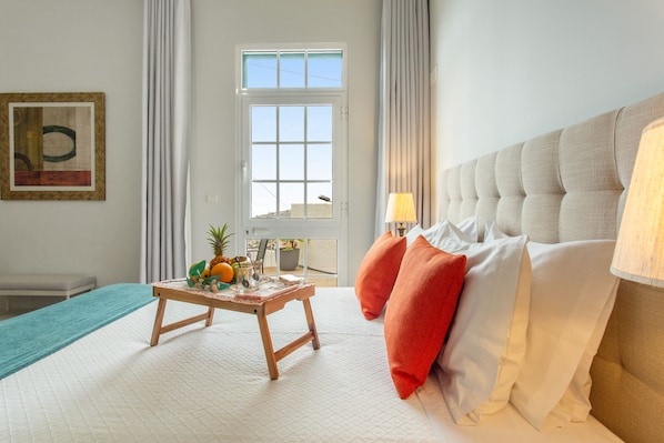 A hotel room with a bed, a small table with fruit, a view through a window, and a painting on the wall. - A Minha Casa – Your Holiday Home in Funchal with Sea Views (Funchal)