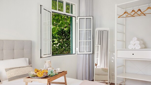 A bedroom with a bed, a wooden side table with fruit and a tray, a wardrobe, and a window with greenery outside.
