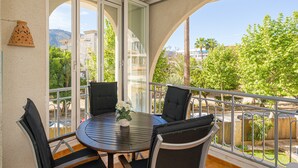 En balkon med et rundt bord og stole, store vinduer og udsigt til grønne områder og bygninger.
