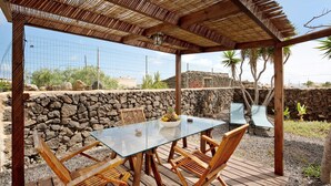 Um pátio ao ar livre com mesa de vidro, cadeiras de madeira, parede de pedra e pérgola.