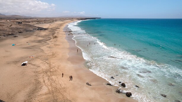 Uma praia com águas azul-claras, areal e algumas pessoas caminhando.