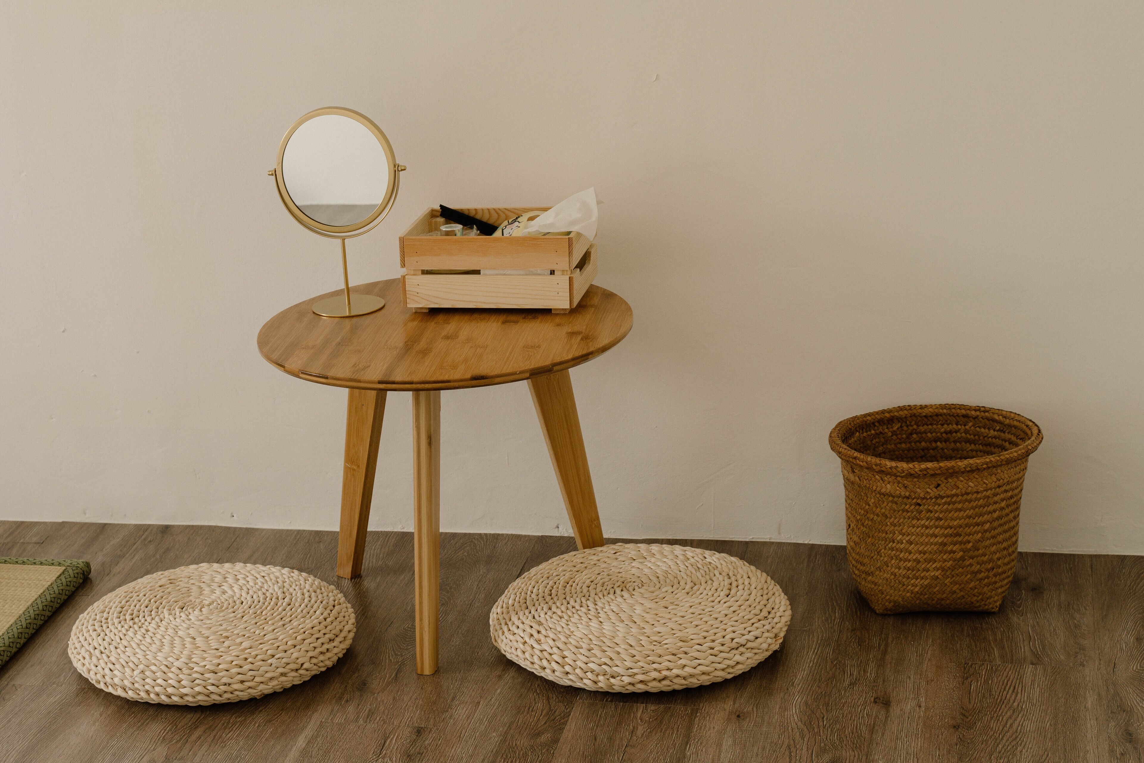 A small wooden table with a round mirror and a box containing tissues. There are two woven poufs and a woven basket on the floor.