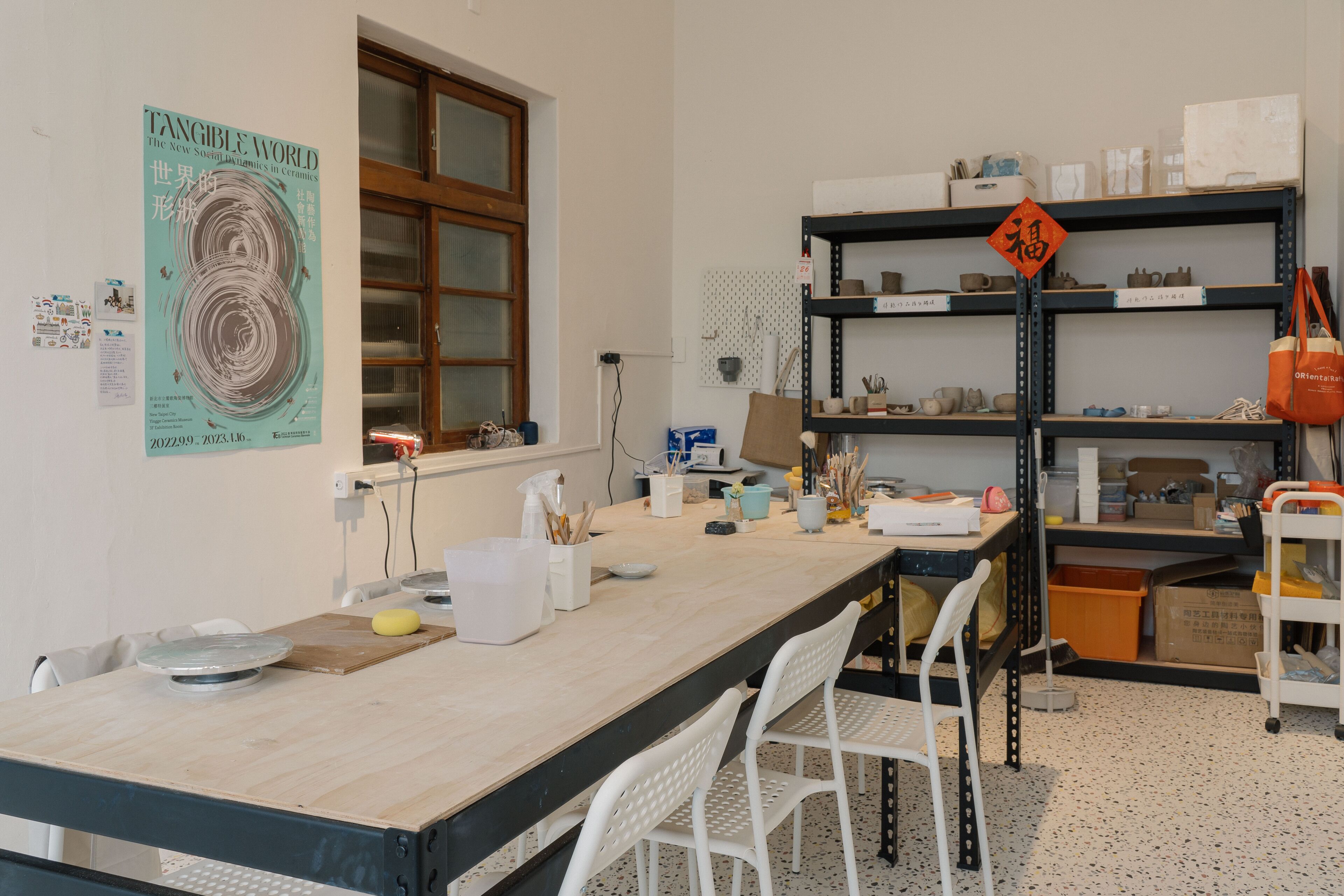 Ein Workshop mit einem langen Tisch, weißen Stühlen, Regalen mit Keramik und einem Poster an der Wand.