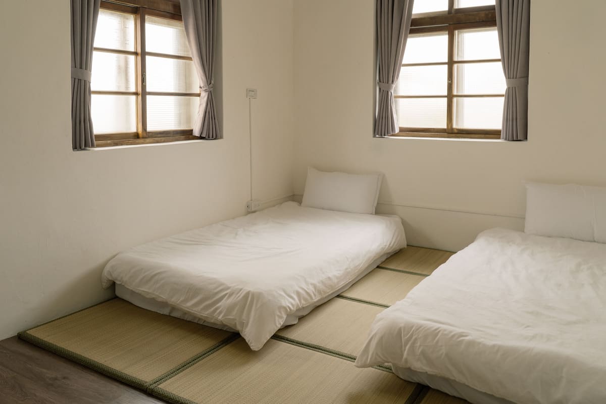 A small, clean room with two single beds, a window with curtains, and a floor mat.
